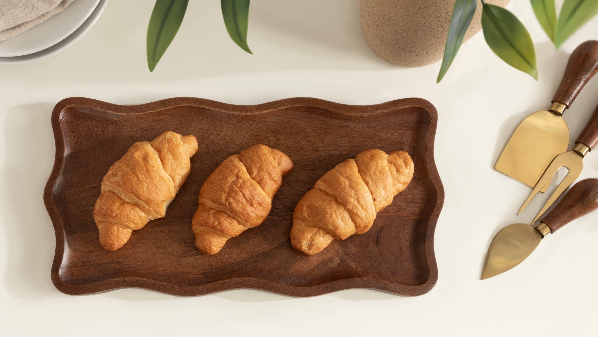 Three croissants on a wooden tray with cheese knives and a glass of milk in the background.