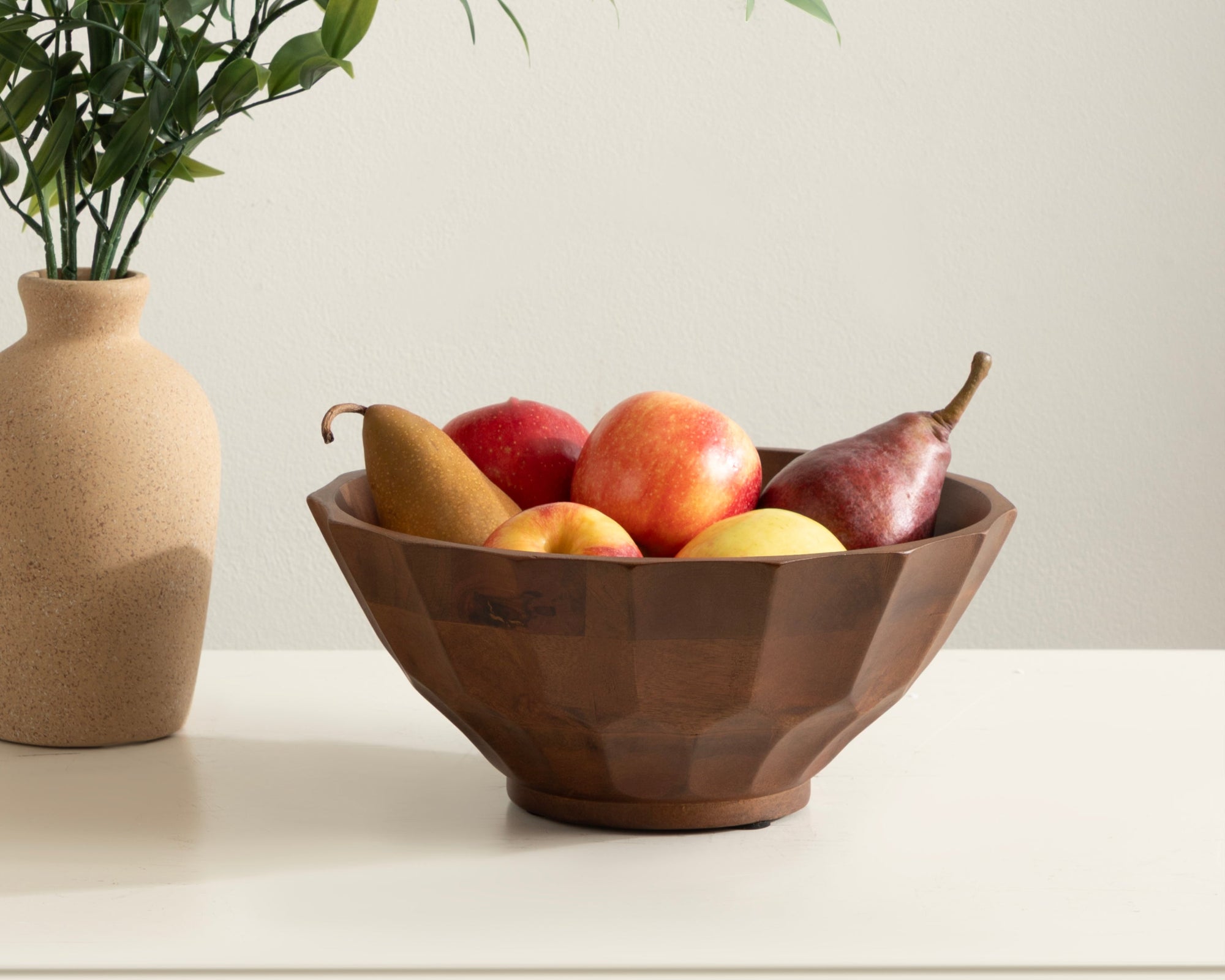 Wooden bowl with fruits on a white surface next to a vase with greenery