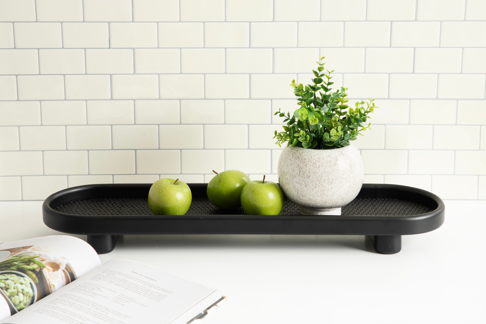 Black tray with green apples and a plant on a white surface with a tiled wall background