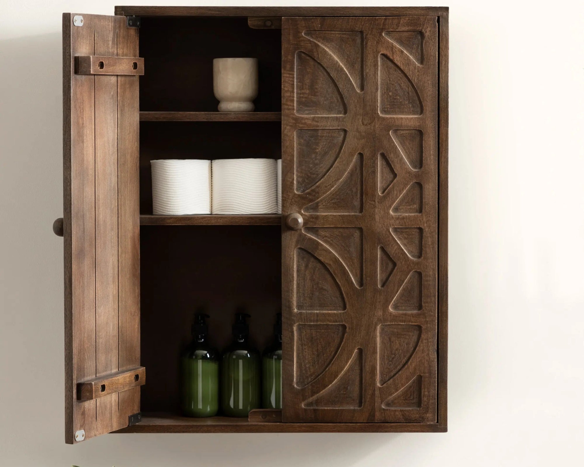 Wooden cabinet with decorative design on a white wall, containing bottles and a plant.
