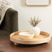 Round wooden tray with a white vase and bowl on a wooden table in a living room setting.