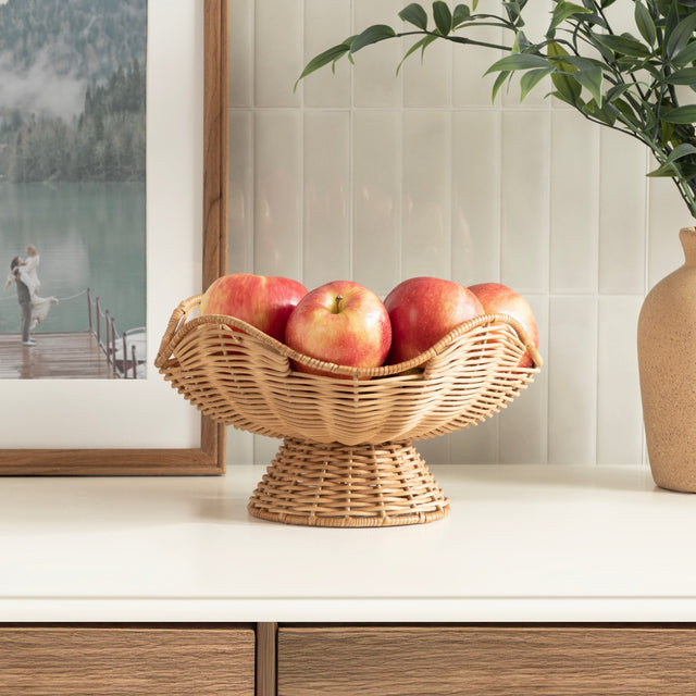 Wicker fruit bowl with apples on a kitchen counter, with a plant and framed picture in the background.