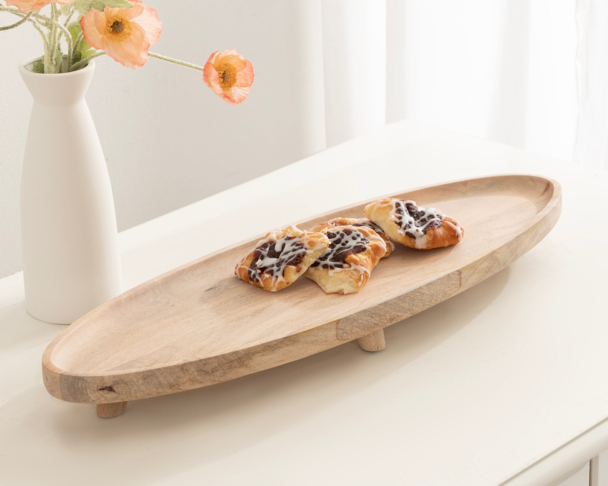 Wooden serving board with pastries on a white surface next to a vase of flowers.